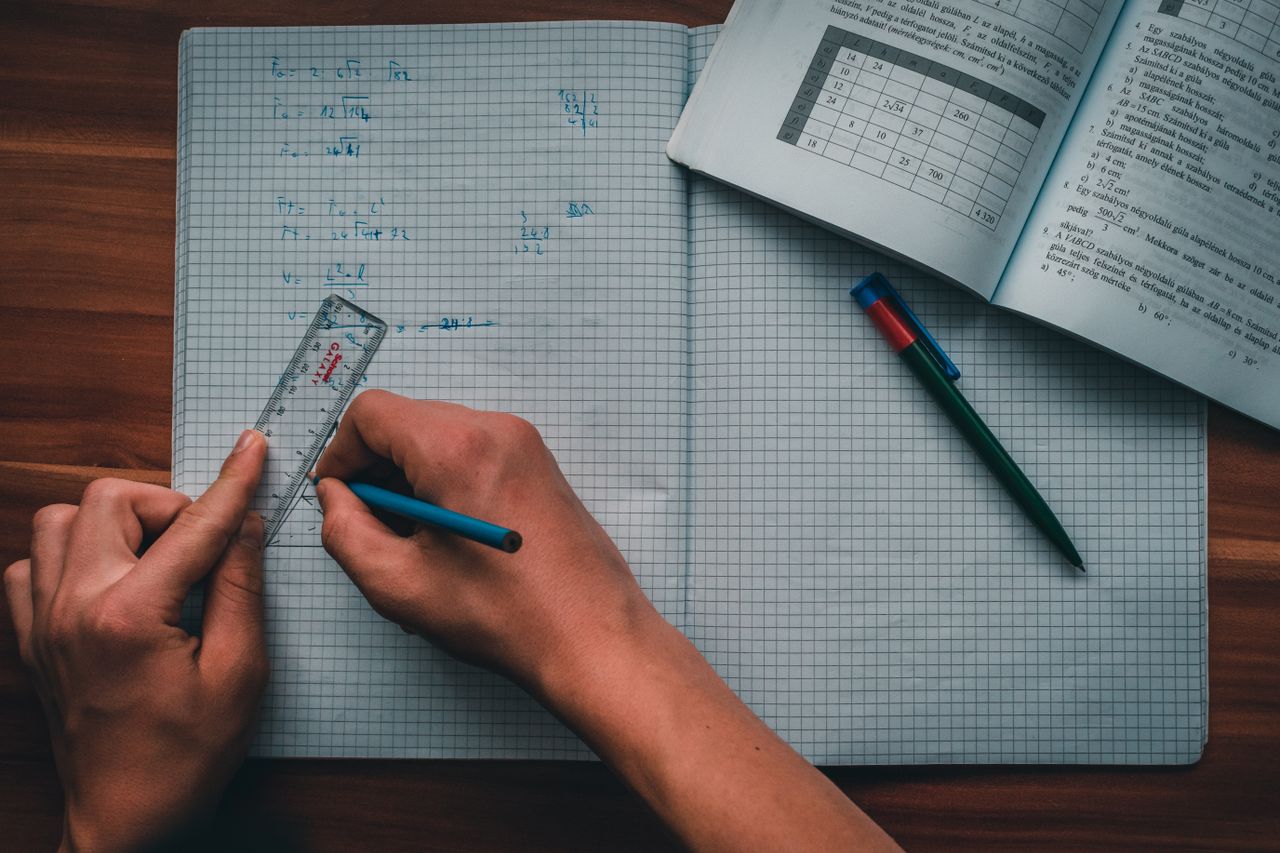 gallery-06 A person writes mathematical equations in a notebook using a ruler and pencil, focused on solving problems.