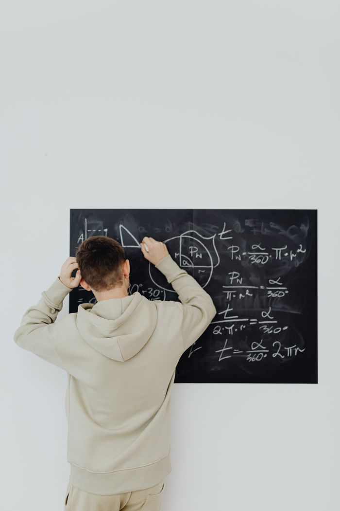 gallery-02 Back view of a teenage boy in a hoodie solving complex math equations on a blackboard indoors.