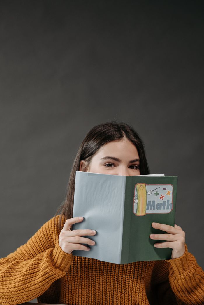 hero A teenage girl holds a math book while partially hidden, highlighted against a gray background.
